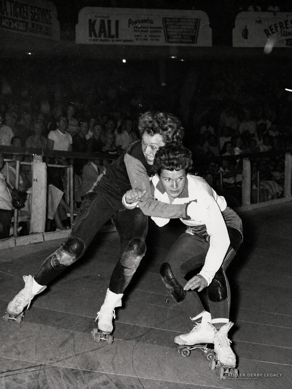 Roller Skaters Grapple on Indoor Track Beneath KALI Sign