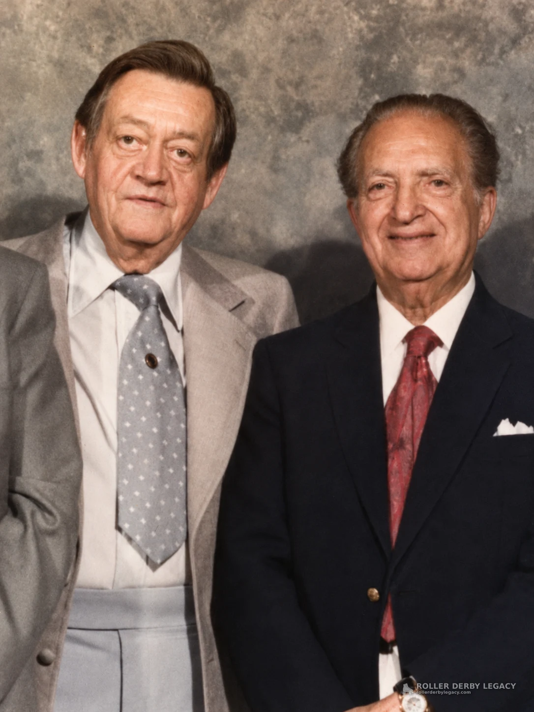 Two older men in formal suits pose side by side against a gray mottled studio backdrop.