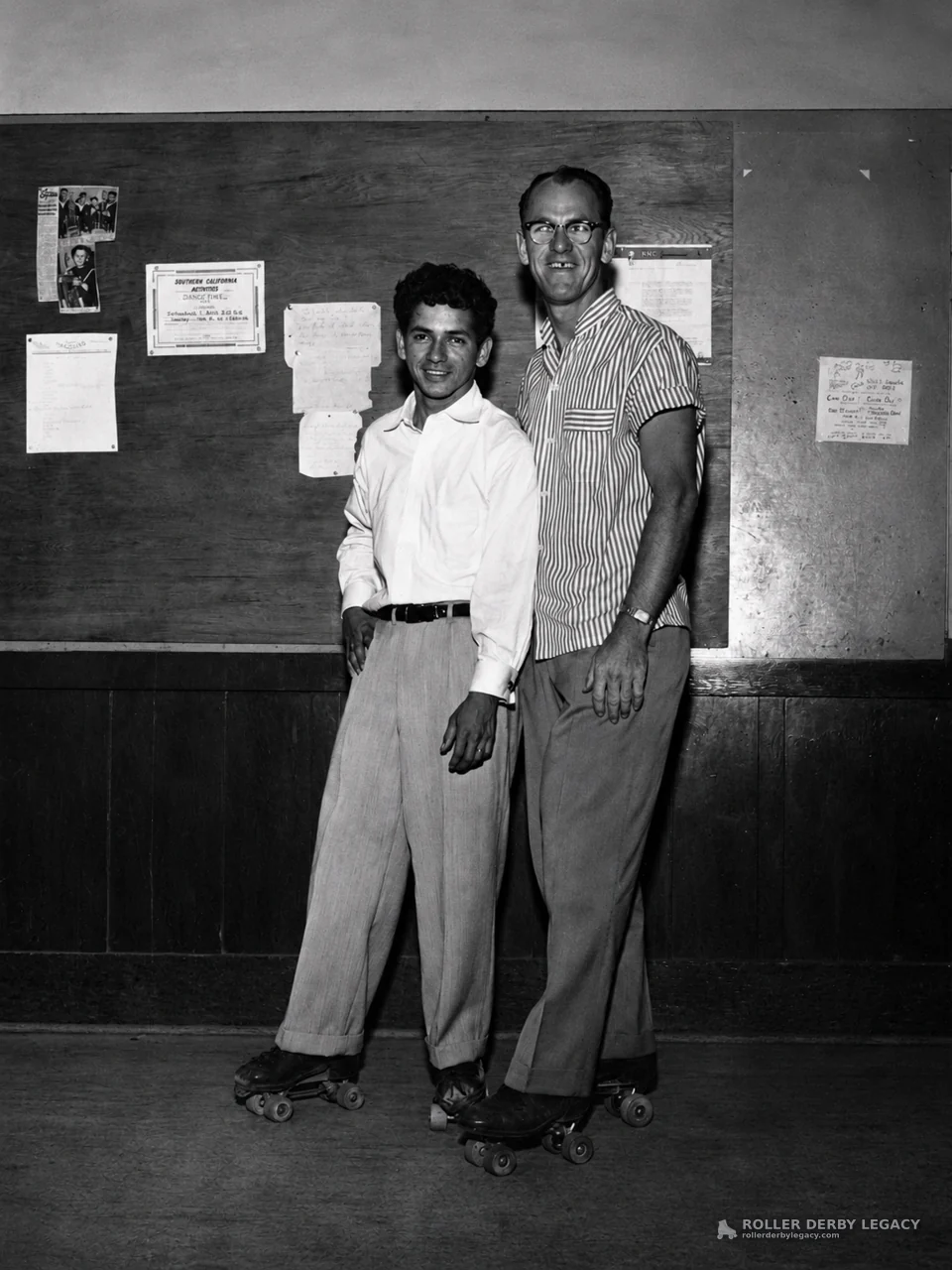 Two Men on Roller Skates Beside a Bulletin Board