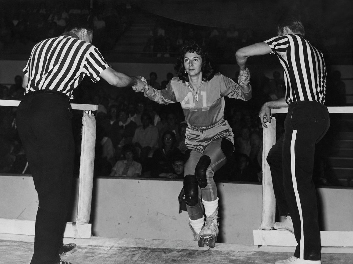 Black-and-white photograph of a roller derby skater wearing jersey number 41 on the banked track, flanked by referees, mid-twentieth century.