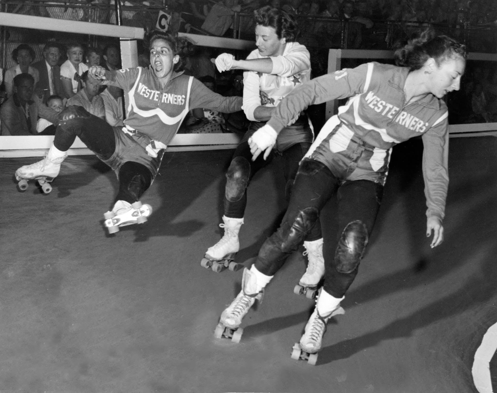 Black-and-white action photograph of four roller derby skaters at the track edge with a large seated crowd watching, mid-twentieth century; one skater is falling at the right side of the frame.